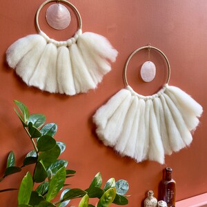 May include: Two white wool wall hangings with a circular shape and a pink shell hanging in the center of each. The hangings are on a terracotta wall.