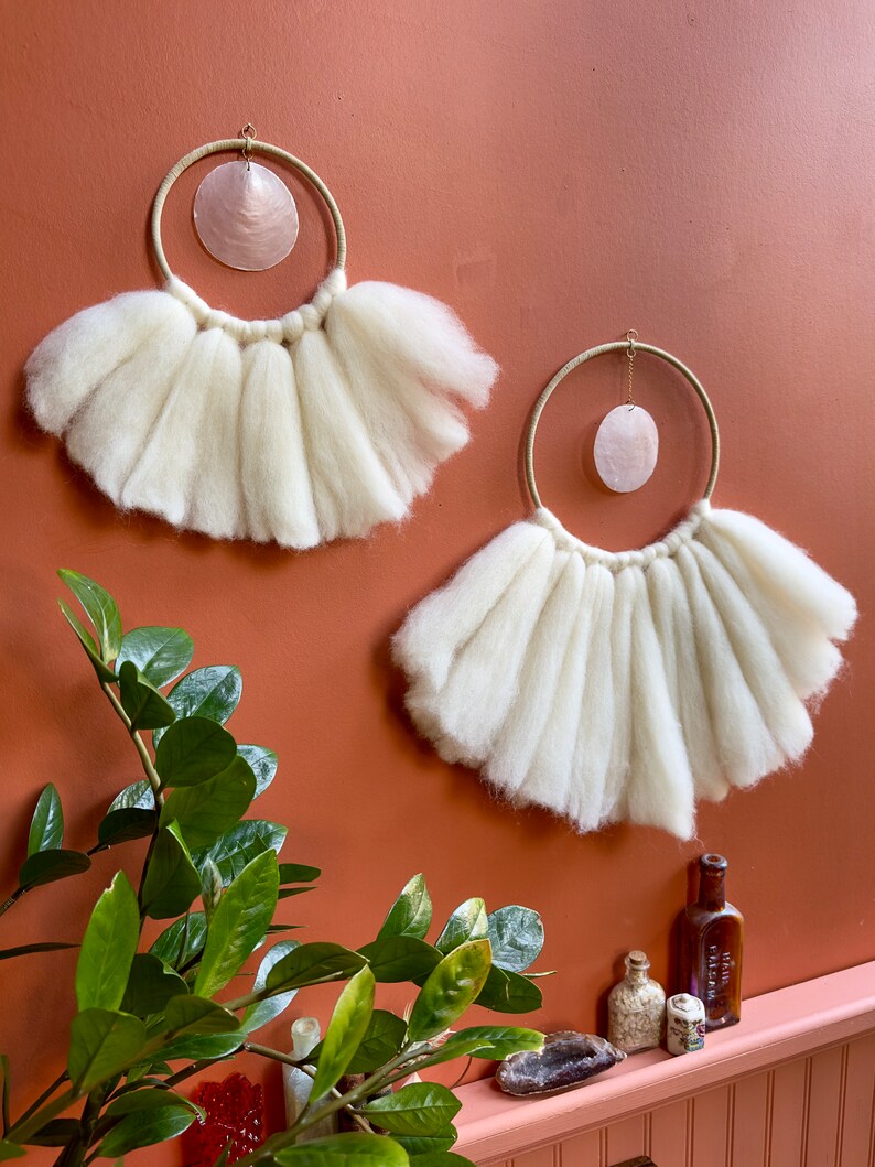 May include: Two white wool wall hangings with a circular shape and a pink shell hanging in the center of each. The hangings are on a terracotta wall.