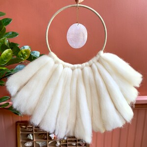 May include: A white wool wall hanging with a large, round, cream-colored hoop and a pink oval-shaped crystal hanging from the hoop.