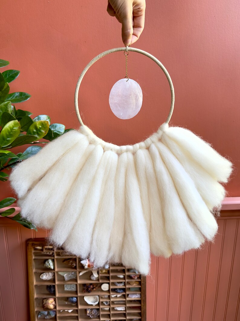 May include: A white wool wall hanging with a large, round, cream-colored hoop and a pink oval-shaped crystal hanging from the hoop.