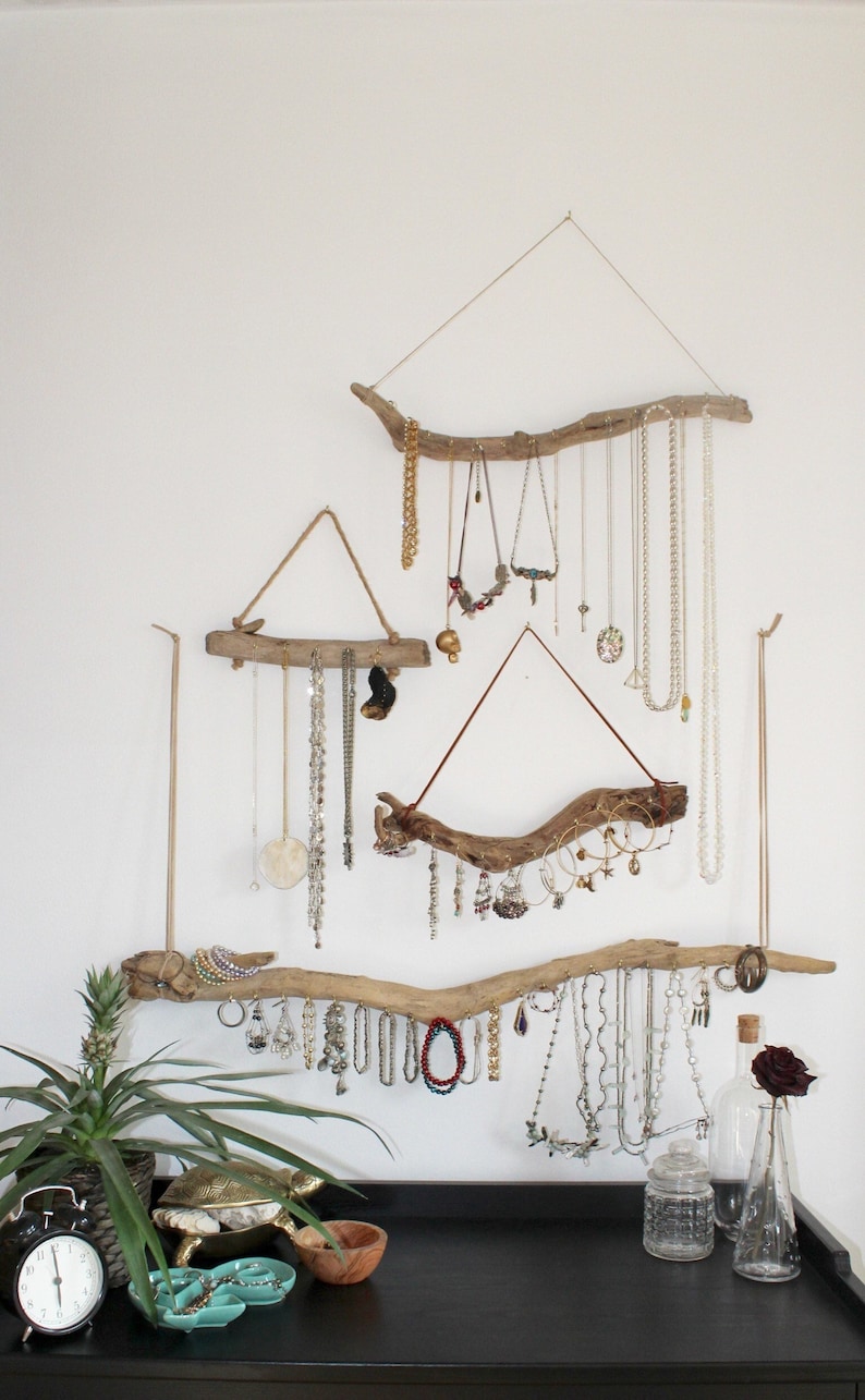 Puede incluir: Un organizador de joyas de pared hecho de ramas de madera flotante. El organizador presenta m&uacute;ltiples ramas de diferentes tama&ntilde;os y formas, cada una con collares, aretes y pulseras colgantes. Las ramas est&aacute;n aseguradas a la pared con cuerda y ganchos.