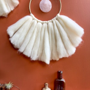 May include: White wool yarn wall hanging with a large, round, pink shell hanging from a wooden hoop.