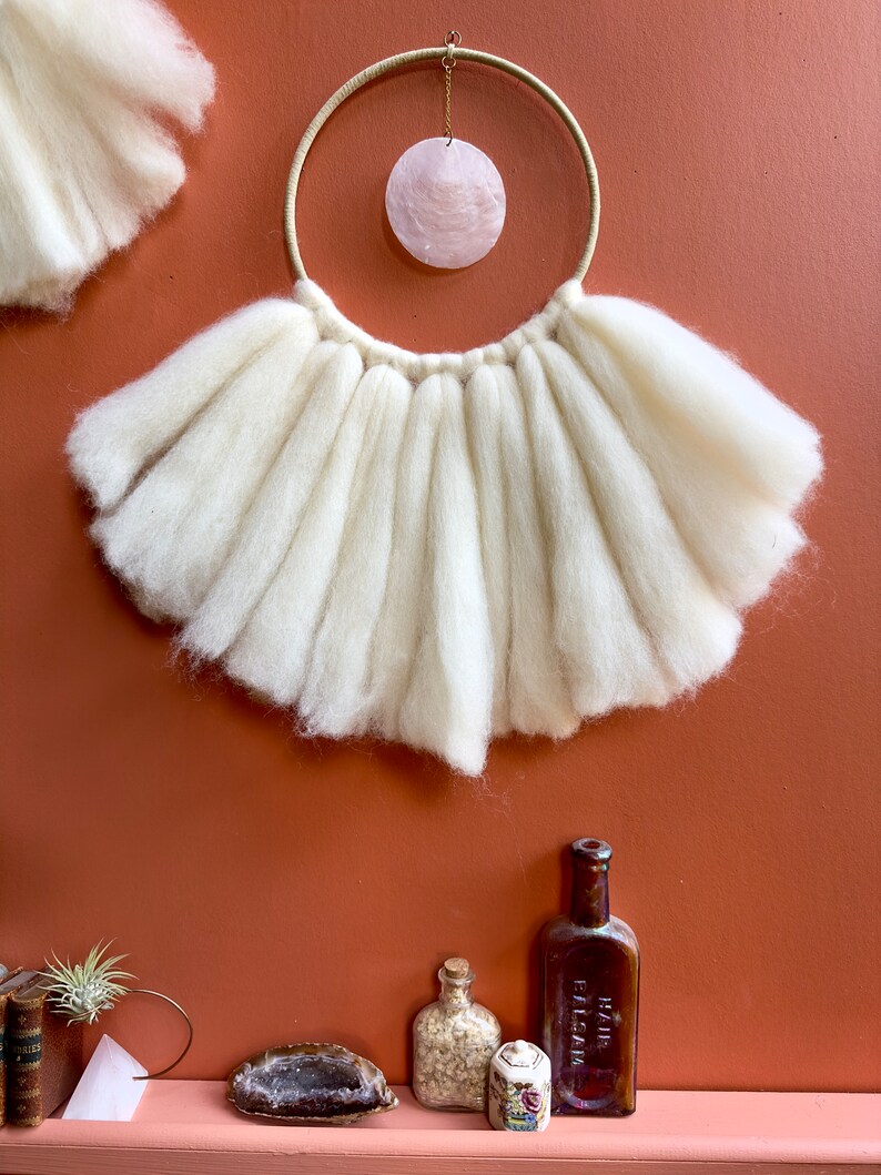 May include: White wool yarn wall hanging with a large, round, pink shell hanging from a wooden hoop.