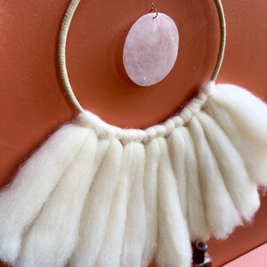 May include: A white macrame wall hanging with a large, round, pink shell hanging from the center. The macrame is made of thick, fluffy yarn and has a fringe around the bottom.