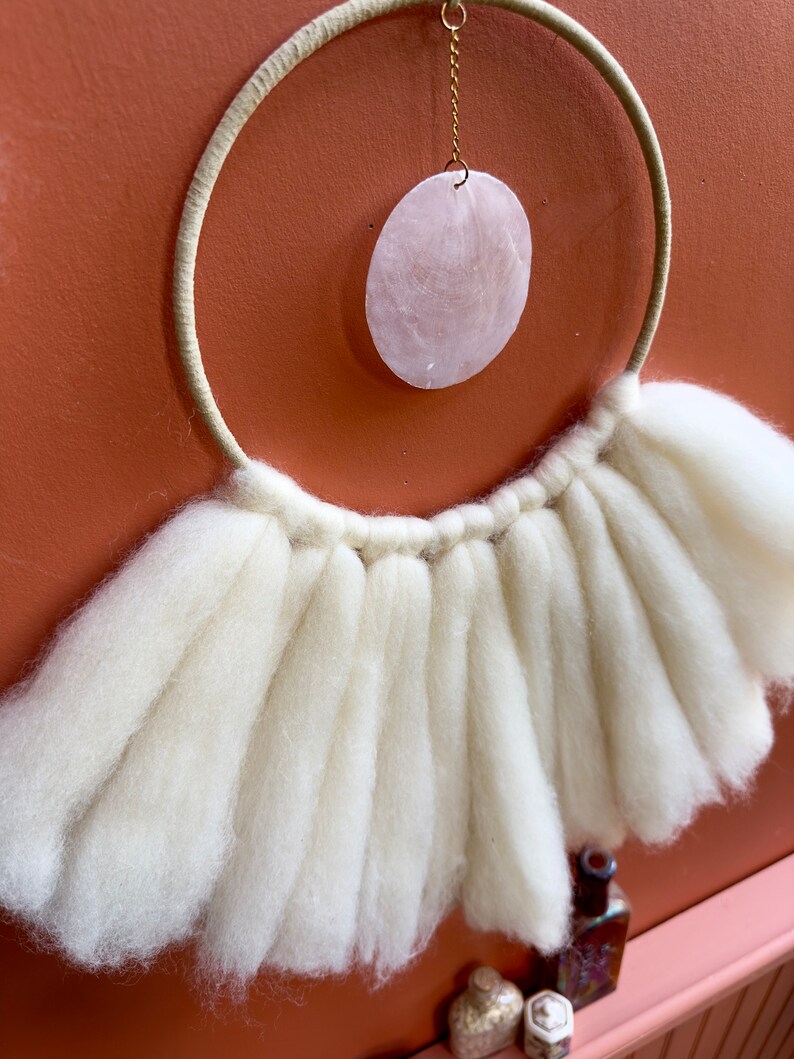 May include: A white macrame wall hanging with a large, round, pink shell hanging from the center. The macrame is made of thick, fluffy yarn and has a fringe around the bottom.