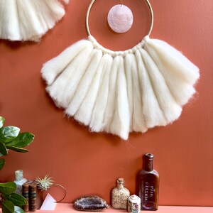 May include: A white wool wall hanging with a large, round, pink shell hanging from a gold hoop. The wall hanging is on a terracotta wall. There are several small bottles and a crystal on a shelf below the wall hanging.