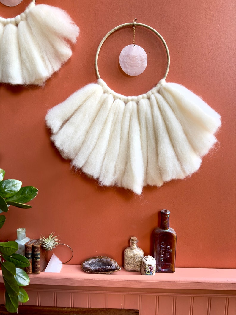 May include: A white wool wall hanging with a large, round, pink shell hanging from a gold hoop. The wall hanging is on a terracotta wall. There are several small bottles and a crystal on a shelf below the wall hanging.