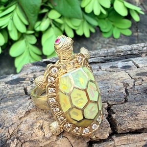 May include: A gold-toned ring shaped like a turtle. The turtle shell features a pale yellow, iridescent inlay with a geometric pattern. The head is accented with a small red stone, and the outer edge of the shell is set with small clear stones.