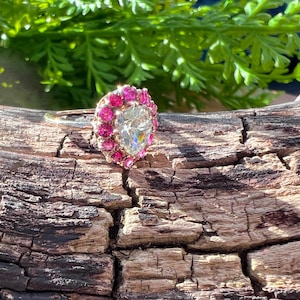 Peut inclure: Une bague en or avec une grosse pierre centrale transparente en forme de cœur, entourée de petites pierres roses. La bague est posée sur une surface en bois brun texturé, avec un feuillage vert en arrière-plan.