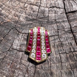 Puede incluir: Broche dorado con diseño geométrico. La broche presenta filas alternas de piedras rojas cuadradas y piedras claras redondas. La pieza está engastada en un marco dorado, creando un contraste llamativo.