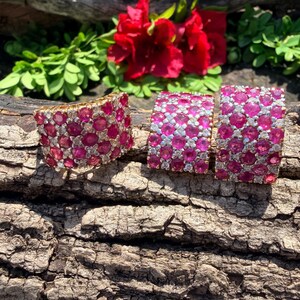 May include: Three rectangular rings encrusted with vibrant pink gemstones and smaller clear stones. The rings are set on a textured, brown wooden surface, with red flowers and green foliage in the background. The rings are a luxurious jewelry item.