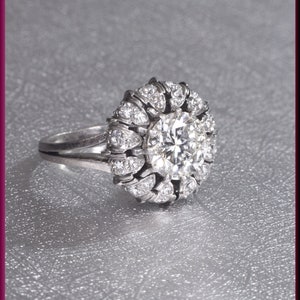 May include: A silver ring with a large, round, clear diamond surrounded by smaller diamonds in a flower-like design.