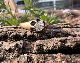 Puede incluir: Un anillo de oro con un solo diamante redondo engastado en garras. El anillo está sobre una pieza de madera.