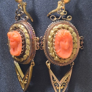 May include: A pair of antique gold earrings with a coral cameo of a woman's face. The earrings have a delicate, ornate design with a dangling, pointed drop.