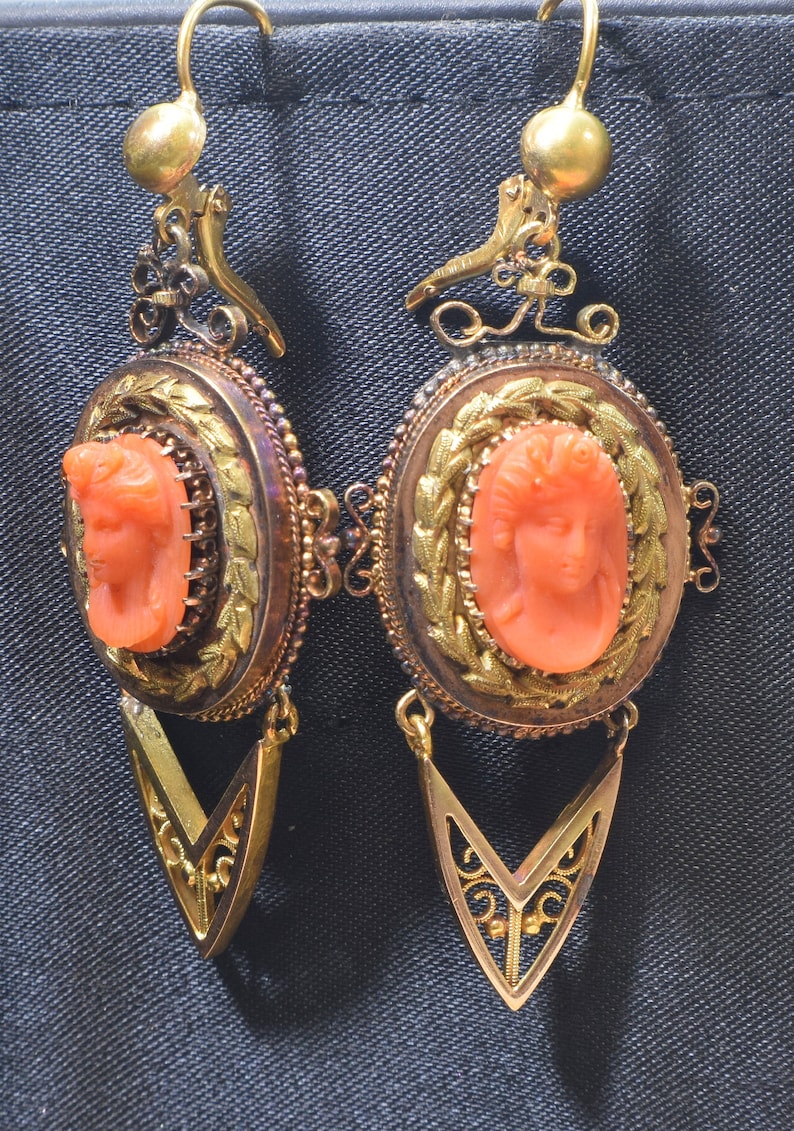 May include: A pair of antique gold earrings with a coral cameo of a woman's face. The earrings have a delicate, ornate design with a dangling, pointed drop.