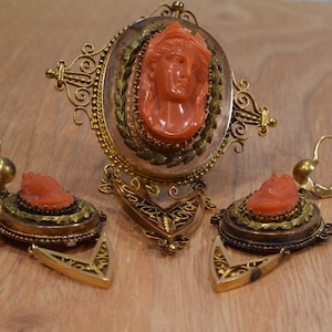 May include: A set of antique coral jewelry featuring a brooch and earrings. The brooch has a cameo of a woman's face carved from coral, surrounded by a gold filigree frame. The earrings are also made of gold and coral, with a smaller cameo of a woman's face on each earring.