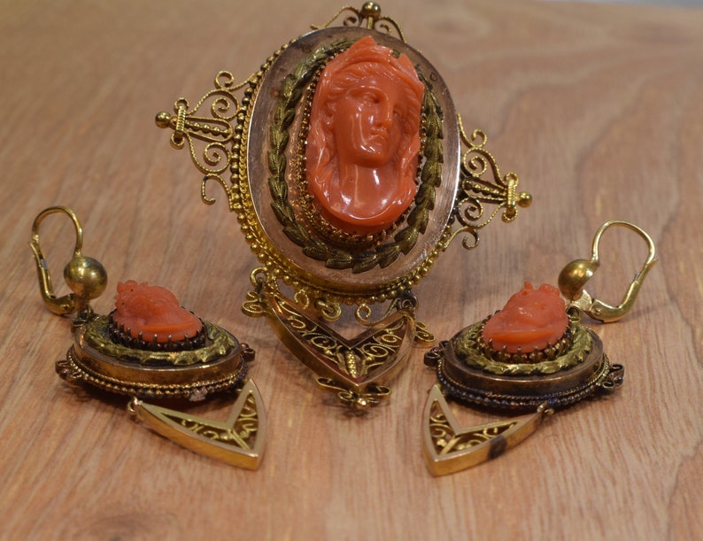 May include: A set of antique coral jewelry featuring a brooch and earrings. The brooch has a cameo of a woman's face carved from coral, surrounded by a gold filigree frame. The earrings are also made of gold and coral, with a smaller cameo of a woman's face on each earring.