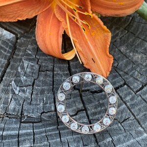 May include: A silver circular brooch with a row of clear round stones around the edge.