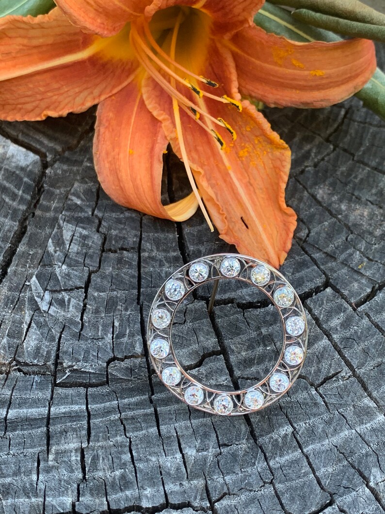 May include: A silver circular brooch with a row of clear round stones around the edge.