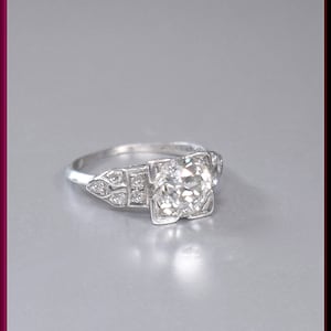 May include: A vintage engagement ring with a large, round, clear diamond set in a platinum band. The diamond is surrounded by smaller diamonds in a delicate, geometric design.