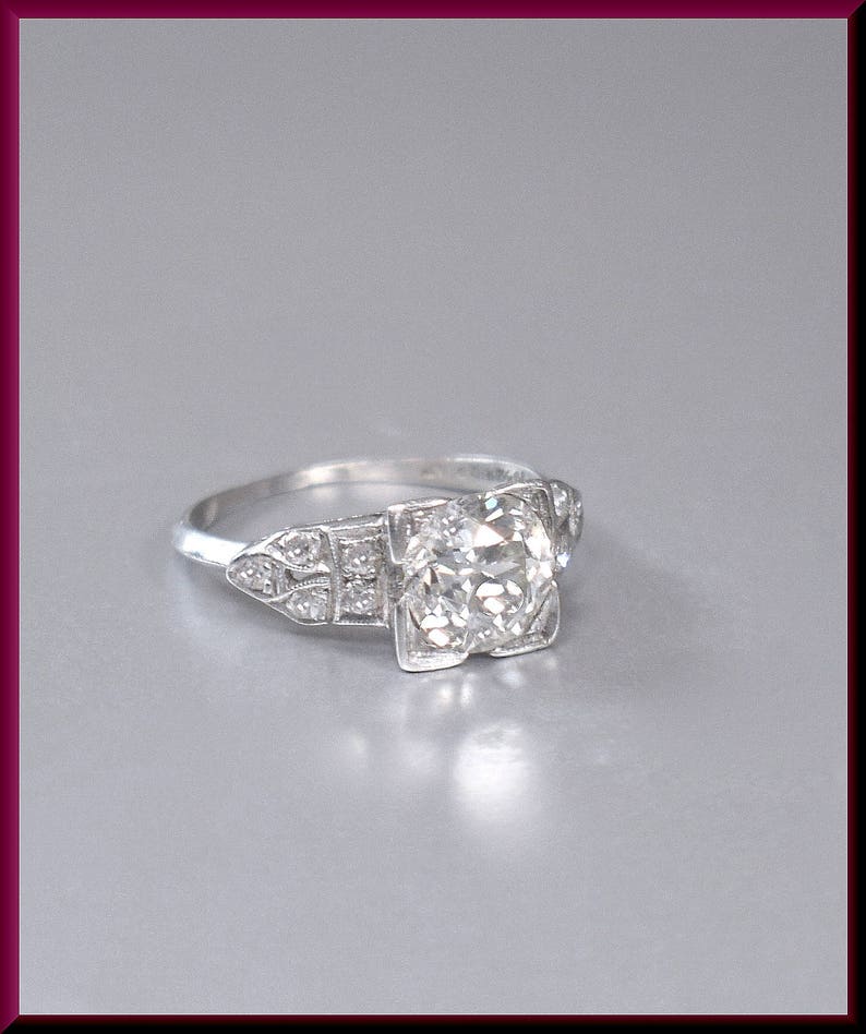 May include: A vintage engagement ring with a large, round, clear diamond set in a platinum band. The diamond is surrounded by smaller diamonds in a delicate, geometric design.