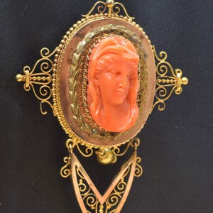 May include: A vintage coral cameo brooch with a gold filigree setting. The cameo features a woman's face carved in detail. The brooch has a dangling pendant with a similar filigree design.