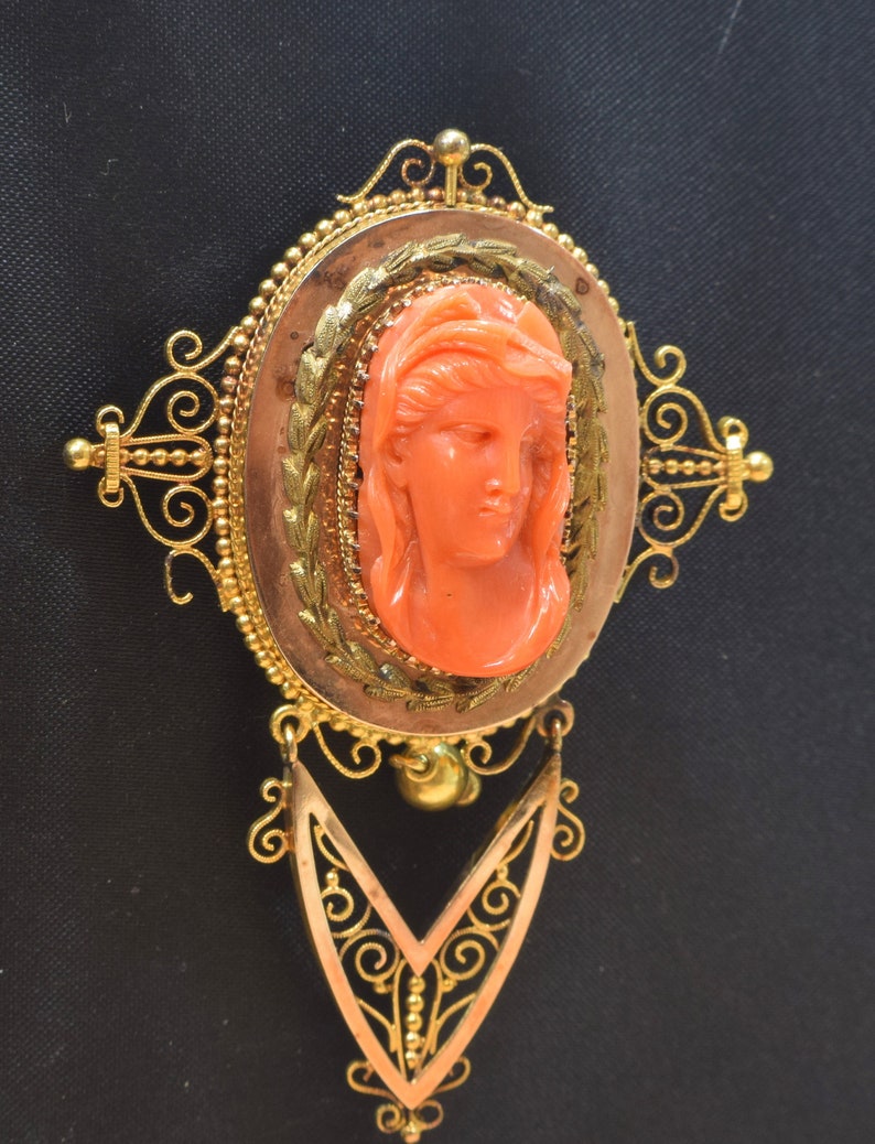 May include: A vintage coral cameo brooch with a gold filigree setting. The cameo features a woman's face carved in detail. The brooch has a dangling pendant with a similar filigree design.
