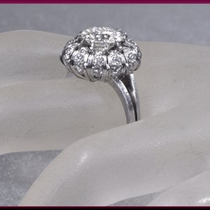 May include: A silver ring with a large, round, diamond-shaped center stone surrounded by smaller diamonds. The ring is set on a white background.