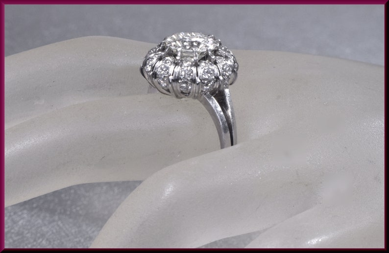 May include: A silver ring with a large, round, diamond-shaped center stone surrounded by smaller diamonds. The ring is set on a white background.