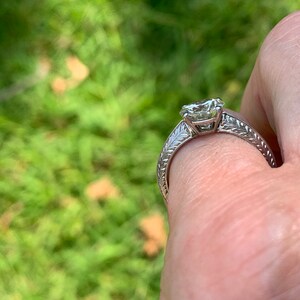 May include: A silver engagement ring with a large, clear, round diamond set in a prong setting. The band is decorated with a delicate, engraved floral design.