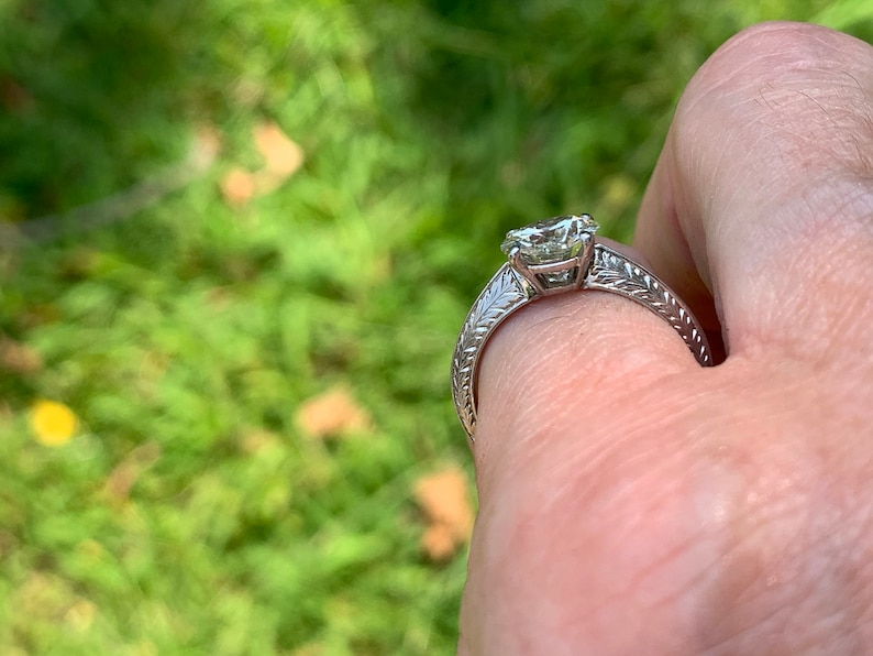May include: A silver engagement ring with a large, clear, round diamond set in a prong setting. The band is decorated with a delicate, engraved floral design.