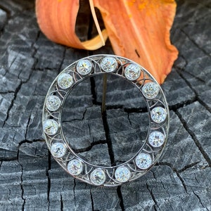 May include: A silver circular brooch with a delicate filigree design and a row of clear round gemstones around the edge.
