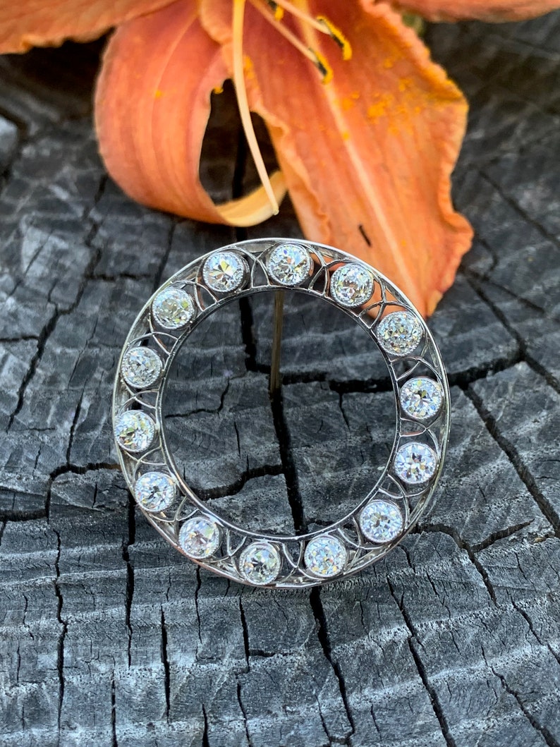 May include: A silver circular brooch with a delicate filigree design and a row of clear round gemstones around the edge.