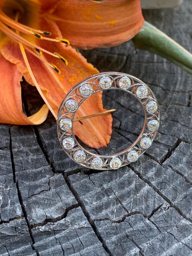 May include: A silver circular brooch with a delicate filigree design and a row of round, clear diamonds around the edge.