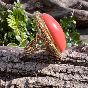 May include: A gold ring featuring a large, oval-shaped, coral gemstone. The ring has an intricate, rope-like design around the stone and band.