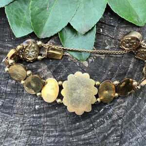 Op de afbeelding: Een gouden armband met een groot, rond, versierd middelpunt en kleinere, ronde, decoratieve schakels. De armband is gemaakt van metaal en heeft een kettingsluiting.