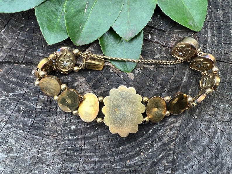 Op de afbeelding: Een gouden armband met een groot, rond, versierd middelpunt en kleinere, ronde, decoratieve schakels. De armband is gemaakt van metaal en heeft een kettingsluiting.