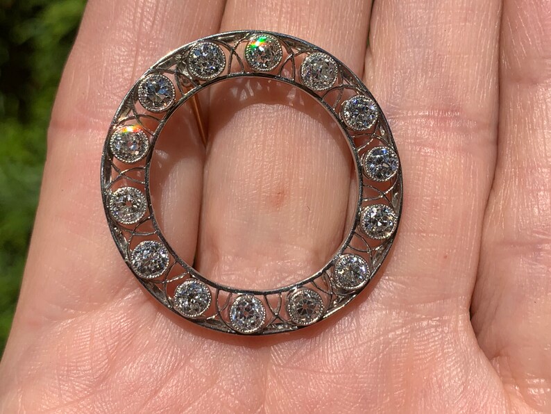 May include: A silver circular brooch with a delicate filigree design and a row of round diamonds around the edge.