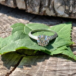May include: A silver engagement ring with a round, clear diamond set in a simple band. The ring is resting on a green leaf.