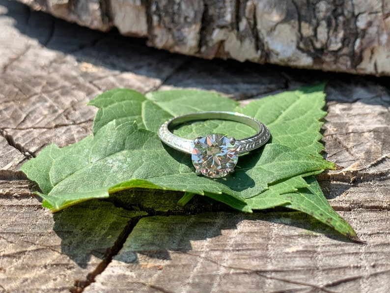 May include: A silver engagement ring with a round, clear diamond set in a simple band. The ring is resting on a green leaf.