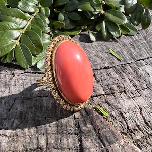 May include: A gold ring featuring a large oval coral stone. The ring is set in a gold band with a rope-like design.