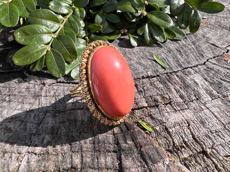 May include: A gold ring featuring a large oval coral stone. The ring is set in a gold band with a rope-like design.