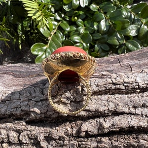May include: A gold ring with a large, round, red gemstone set in a gold bezel. The ring is on a wooden surface.