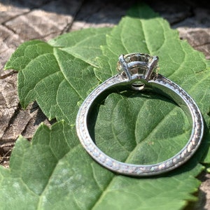 May include: A silver engagement ring with a round diamond set in a prong setting. The band is decorated with a delicate, textured design.