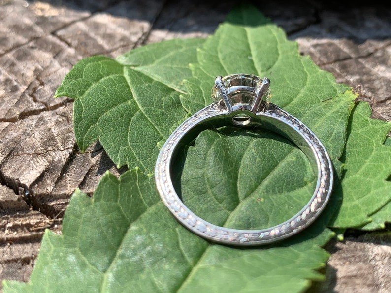May include: A silver engagement ring with a round diamond set in a prong setting. The band is decorated with a delicate, textured design.