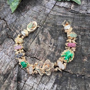 Puede incluir: Una pulsera dorada con motivos de ranas y flores, engastada con piedras preciosas. La pulsera incluye piedras verdes, rojas y azules, y un cierre de broche. Se muestra sobre una superficie de madera envejecida.