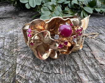 May include: A gold bracelet with a floral design featuring a large red cabochon gemstone surrounded by smaller red gemstones and diamonds.