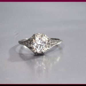 May include: A silver ring with a round, clear diamond set in a filigree design.