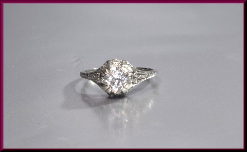 May include: A silver ring with a round, clear diamond set in a filigree design.
