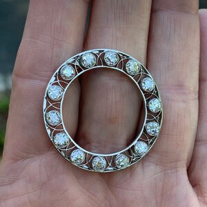 May include: A silver ring with a circular design featuring a row of round diamonds set in a delicate filigree pattern.